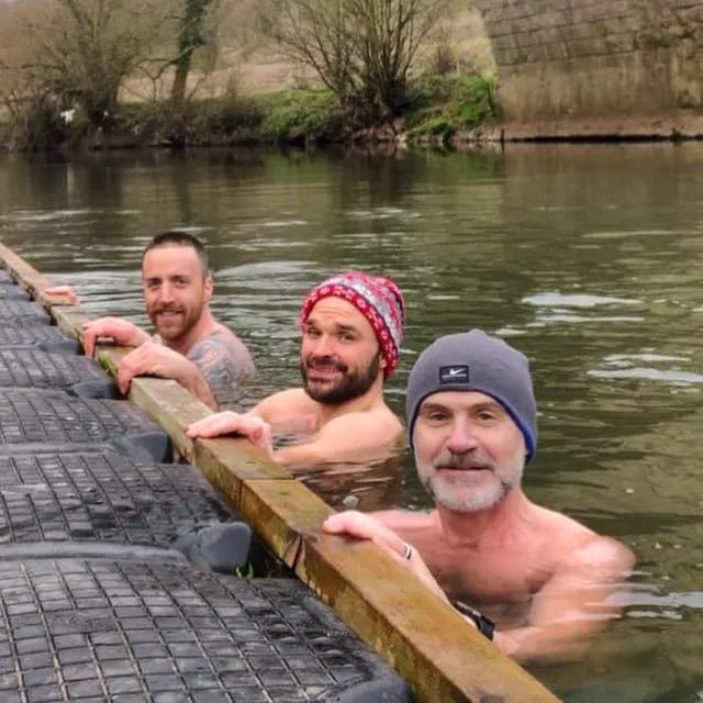 Three of us in a UK river in winter, leaning on the wooden walkway, smiling.